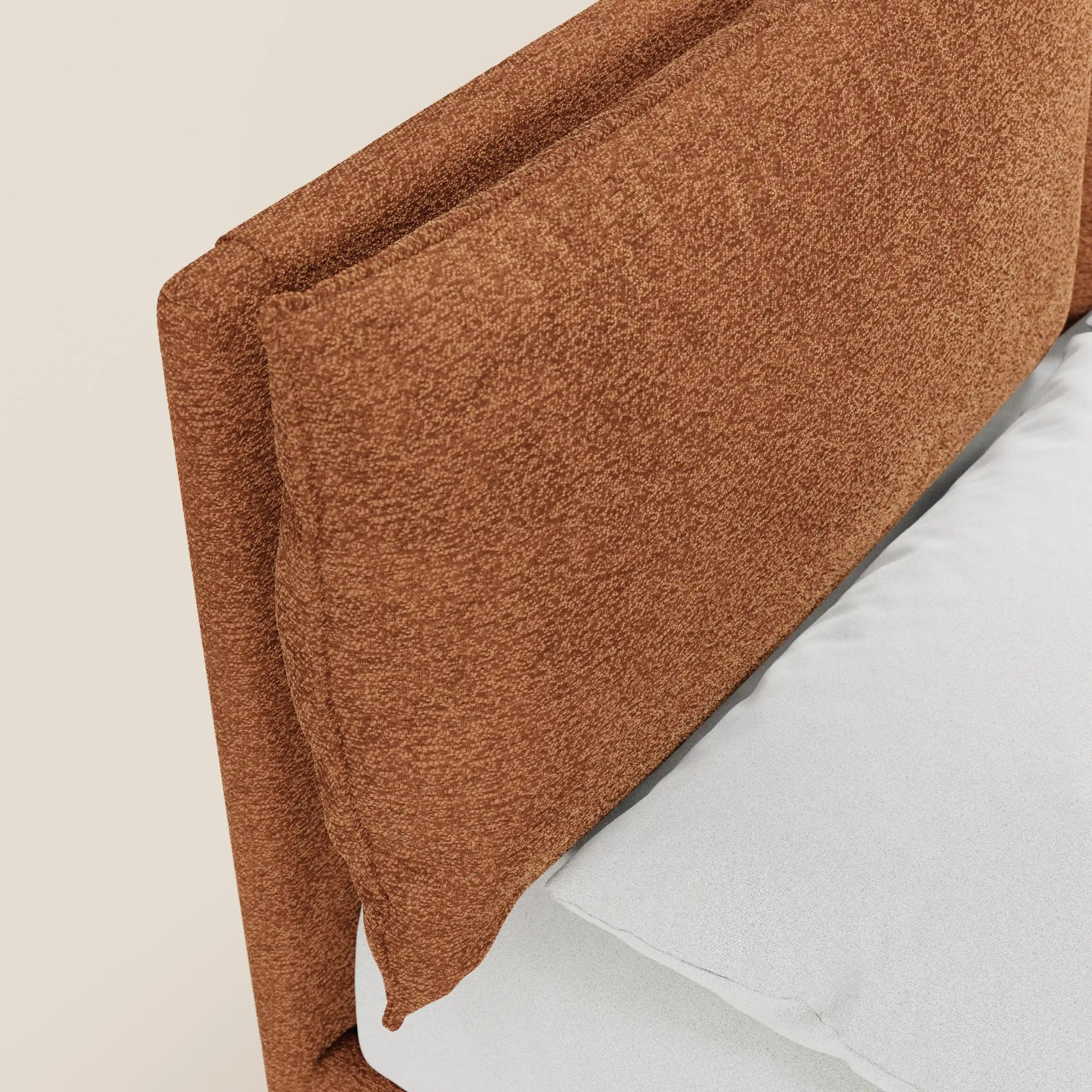 Primo piano del letto imbottito ZAYRON di Letti.store, caratterizzato da un tessuto color ruggine certificato Oeko-Tex®, cuscini imbottiti regolabili e una base contenitore. La testiera risalta sulla parete beige chiaro accanto a un letto ben rifatto con lenzuola bianche.