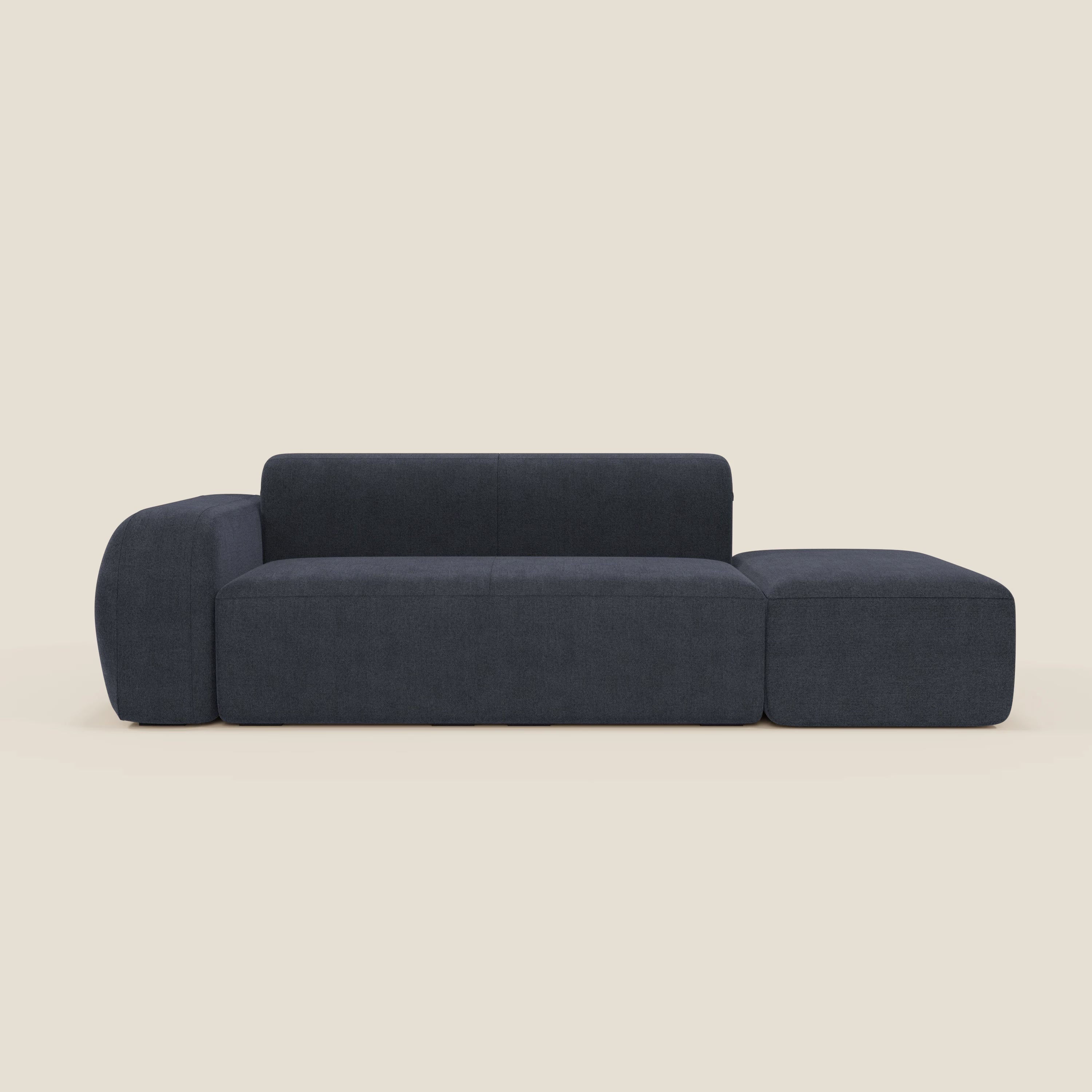 Il BonBon Divano componibile di Divani.Store è caratterizzato da un tessuto blu scuro T23 blu ardesia smacchiabile, schienale basso, ampio bracciolo sinistro e chaise lounge destra con pouf terminale, su uno sfondo beige chiaro.