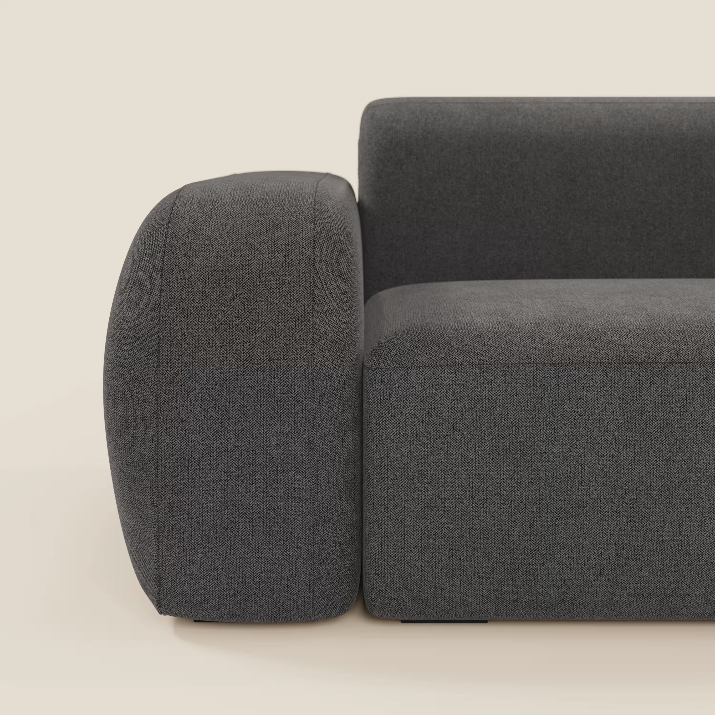 Un primo piano del divano angolare componibile e modulare BonBon di Divani.Store in tessuto smacchiabile T23 antracite mette in evidenza l'ampio bracciolo arrotondato, la seduta e lo schienale, con un'imbottitura strutturata e un design minimalista su uno sfondo beige chiaro.