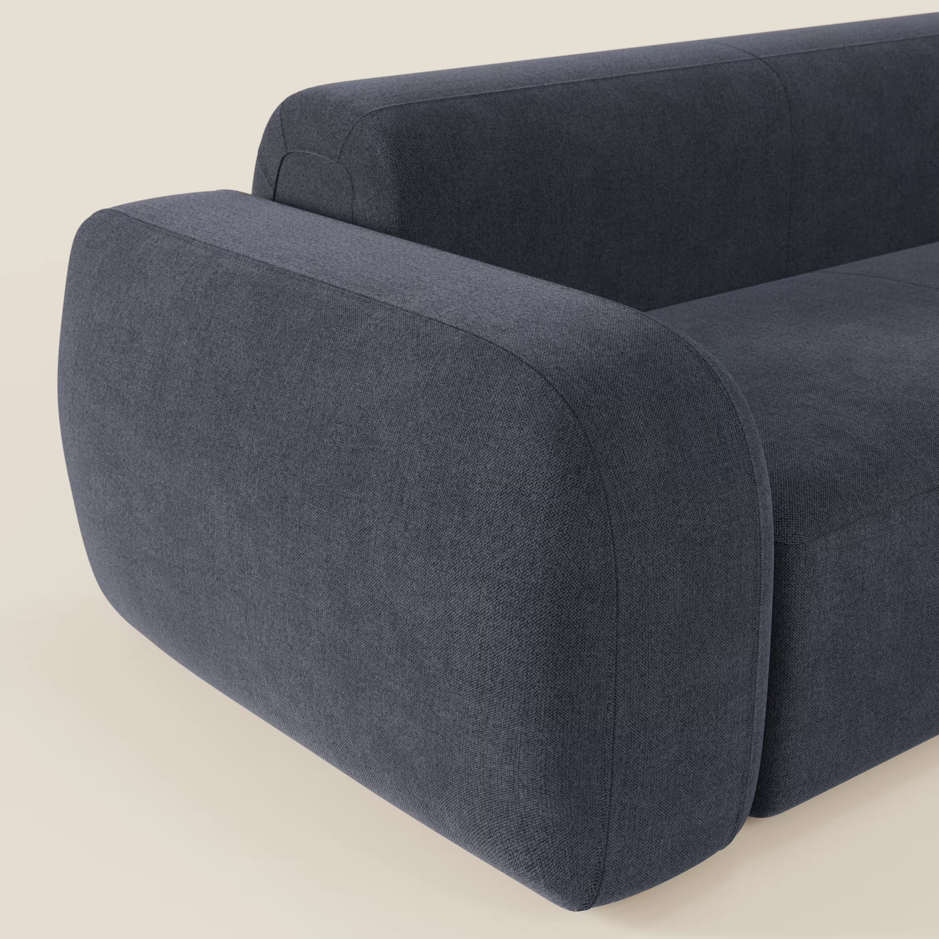 Primo piano del divano angolare componibile e modulare BonBon di Divani.Store, in tessuto smacchiabile T23 blu ardesia. Il divano è caratterizzato da braccioli e schienale ampi e arrotondati, curve morbide e un design minimalista su uno sfondo beige chiaro.