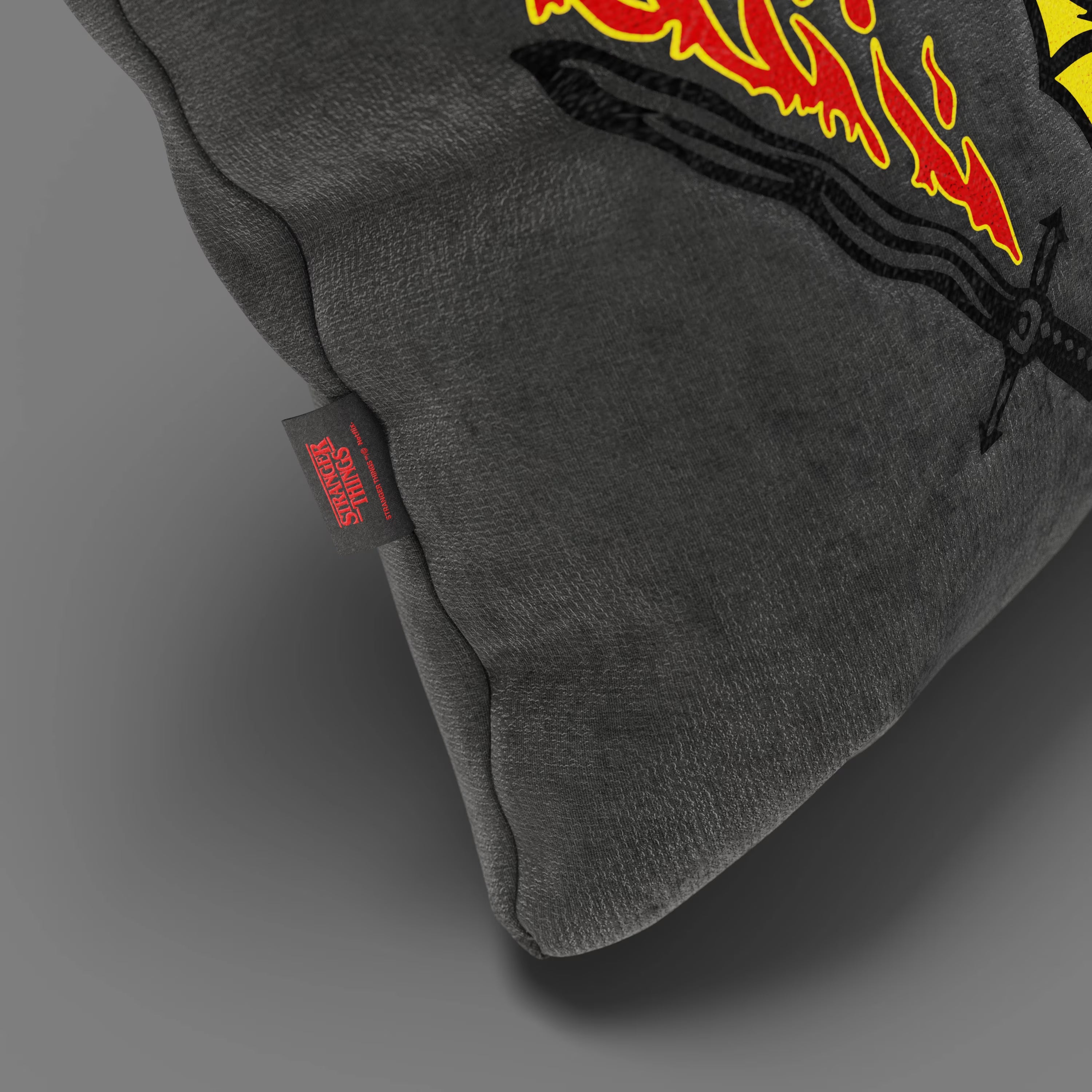 Primo piano del Cuscino Hellfire Club Stranger Things Limited Edition (45x45 cm, antracite) di Divani.Store con fiamme e spada ricamate, con etichetta rossa "Stranger Things". Realizzato in tessuto antimacchia. Fondo grigio liscio.