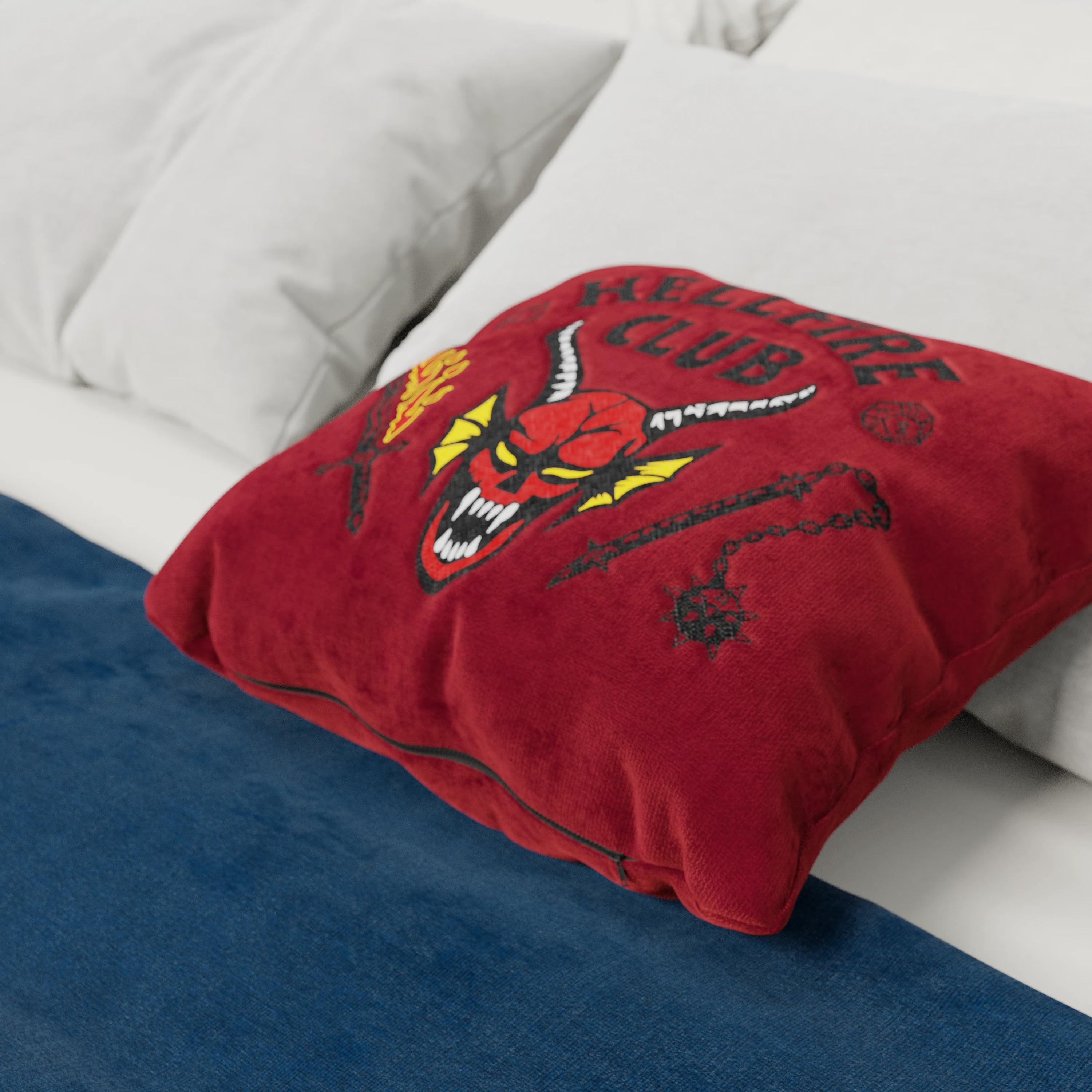 Sul letto c'è un cuscino Divani.Store Hellfire Club Stranger Things Limited Edition rosso 45x45 con corna ricamate, zanne, torcia e la scritta "HELLFIRE CLUB". Il letto ha anche cuscini bianchi e una coperta blu.