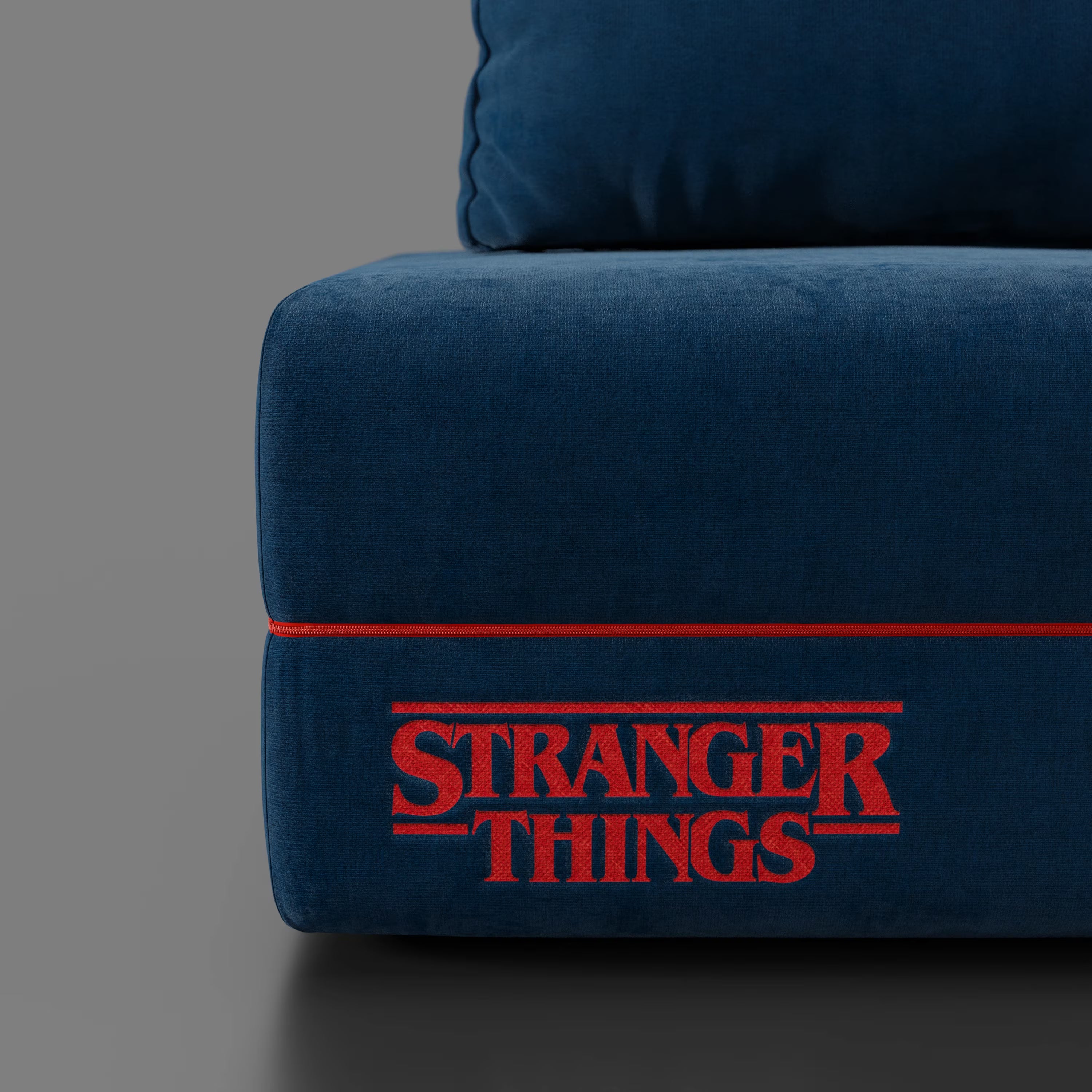 Primo piano della poltrona letto STRANGER THINGS Edition di Divani.Store, con tessuto blu antimacchia e materasso in memory foam da 22 cm. Il logo rosso di "Stranger Things" risalta sul cuscino contro lo sfondo grigio.