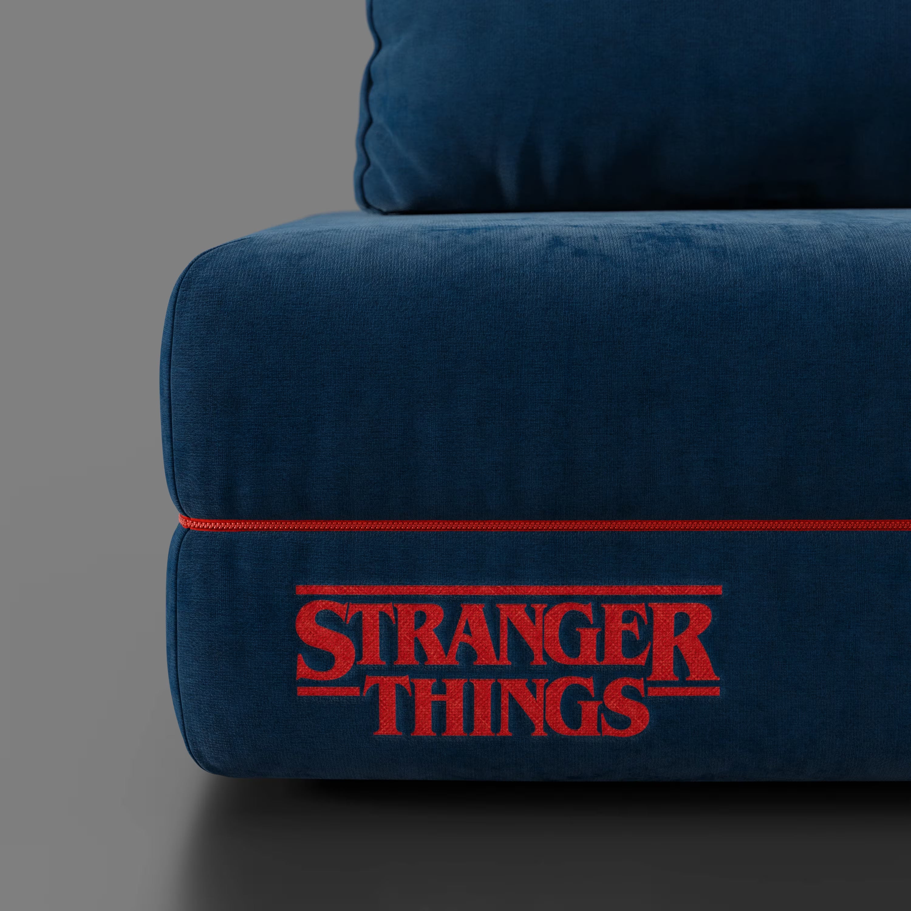 Un primo piano del divano letto matrimoniale Rift STRANGER THINGS Edition di Divani.Store in blu smacchiabile con profili rossi. Il cuscino inferiore mostra il logo "Stranger Things" ricamato in rosso. Lo sfondo è grigio semplice.