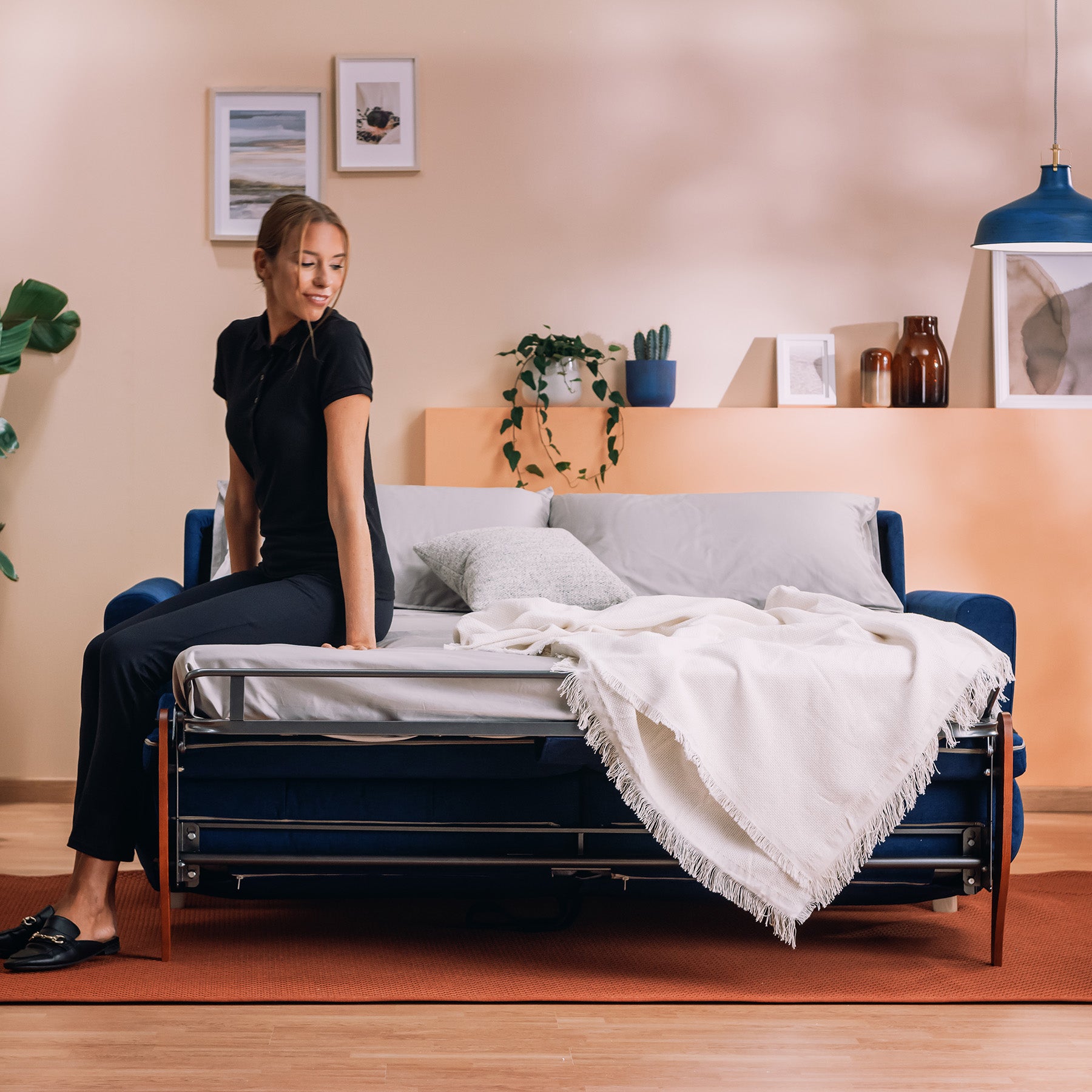 Una donna in nero siede sul letto matrimoniale Callisto Divano in tessuto morbido impermeabile T02 blu di Divani.Store, abbinato a biancheria da letto bianca e a una coperta leggera. L'accogliente stanza ha un'illuminazione calda, opere d'arte incorniciate, piante e scaffali con oggetti decorativi.