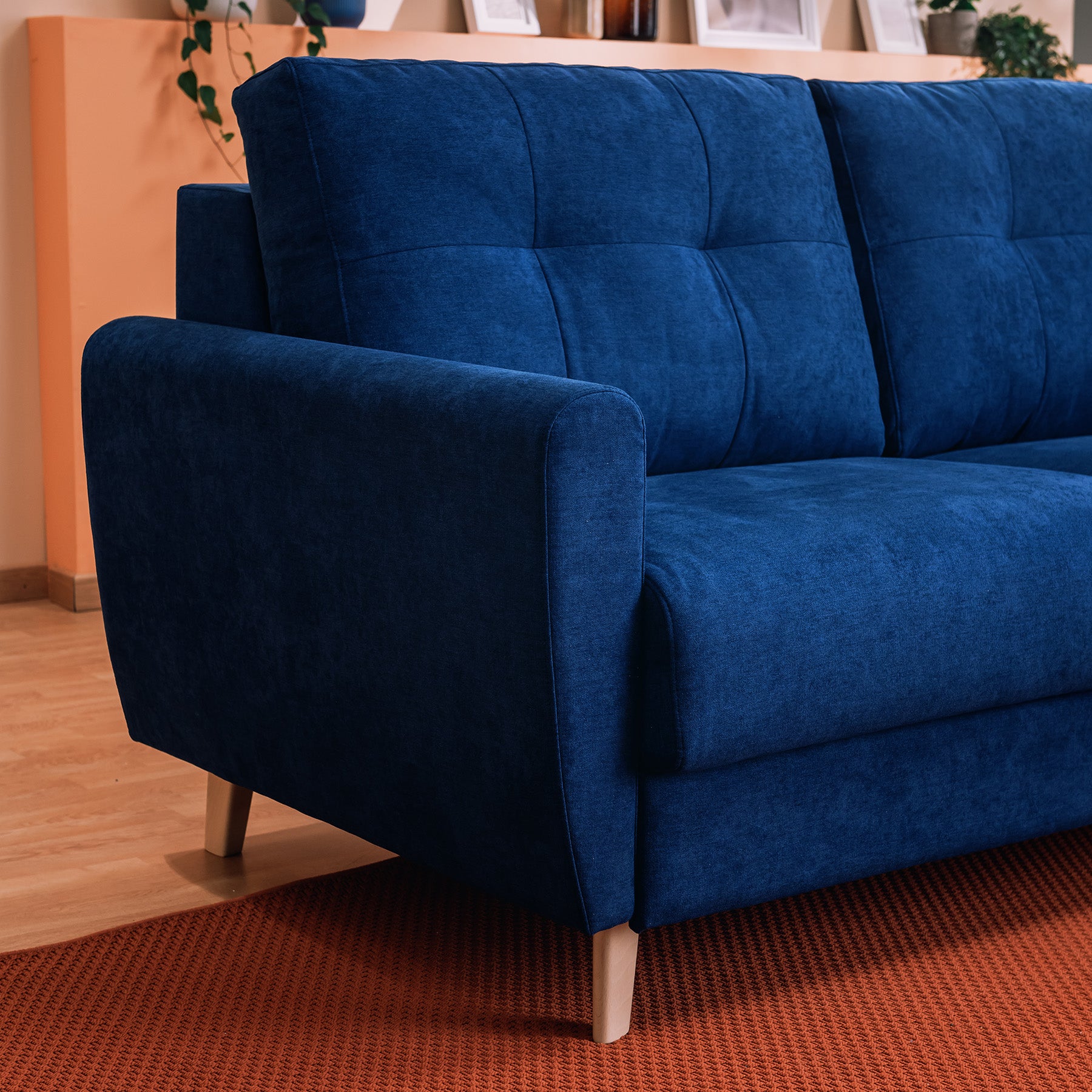 Il Callisto Divano letto matrimoniale in tessuto morbido impermeabile T02 blu di Divani.Store, con gambe in legno e schienale trapuntato, poggia su un tappeto arancione in un soggiorno con pavimento in legno chiaro, piante e libri visibili su una mensola vicina.