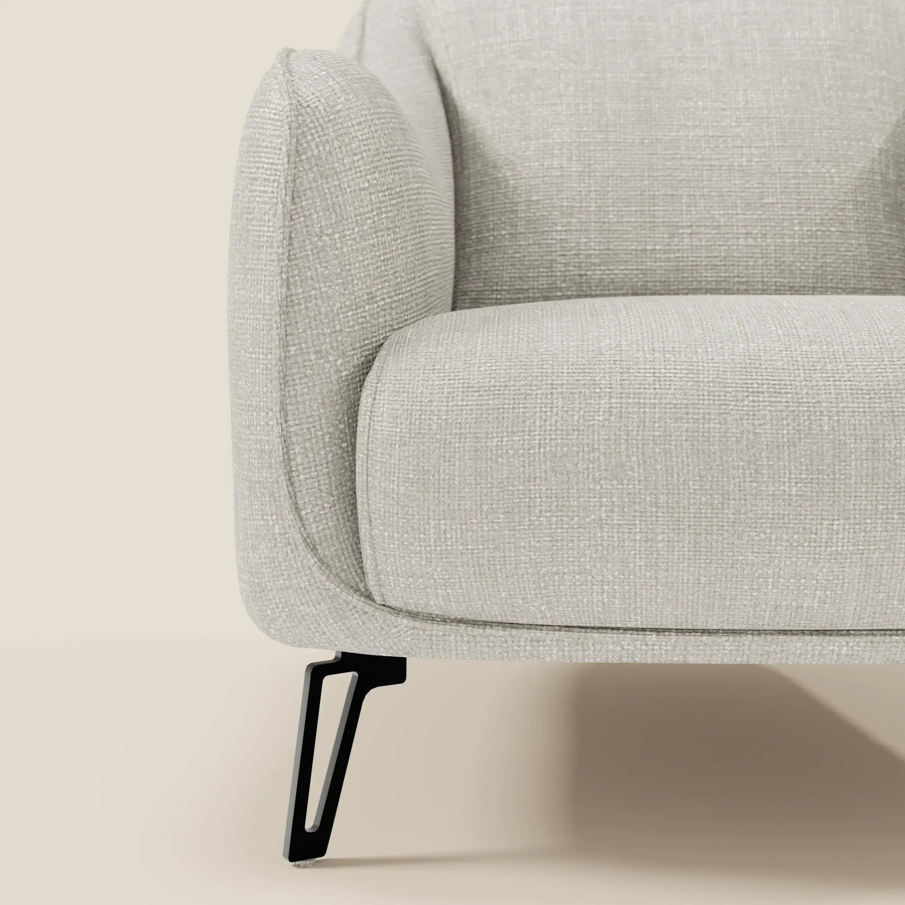 Primo piano della poltroncina Avela di Divani.Store in morbido tessuto impermeabile T06 grigio chiaro, con bracciolo arrotondato e gamba in metallo nero con disegno geometrico aperto. Stile moderno su sfondo beige.