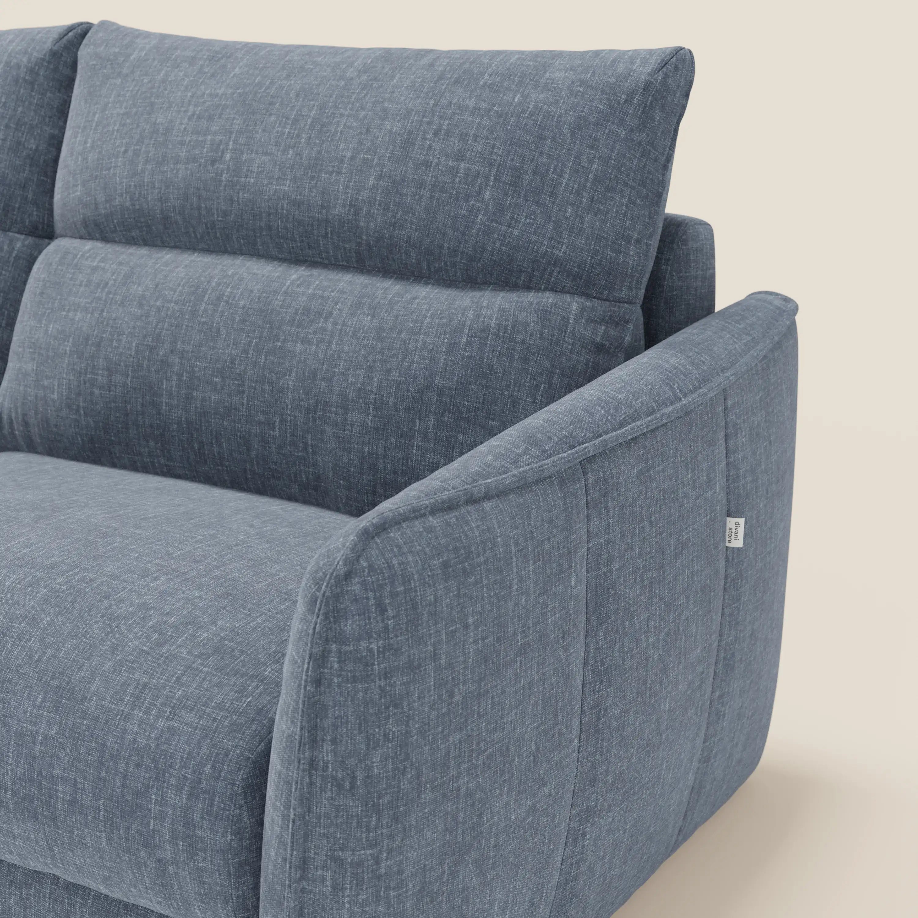 Primo piano del Divano letto Elios di Divani.Store, caratterizzato da cuscini di schienale e braccioli imbottiti e delicatamente inclinati. Rivestito in ardesia blu T24 tessuto smacchiabile. Una piccola etichetta in tessuto bianco sottolinea la cucitura laterale su uno sfondo beige.
