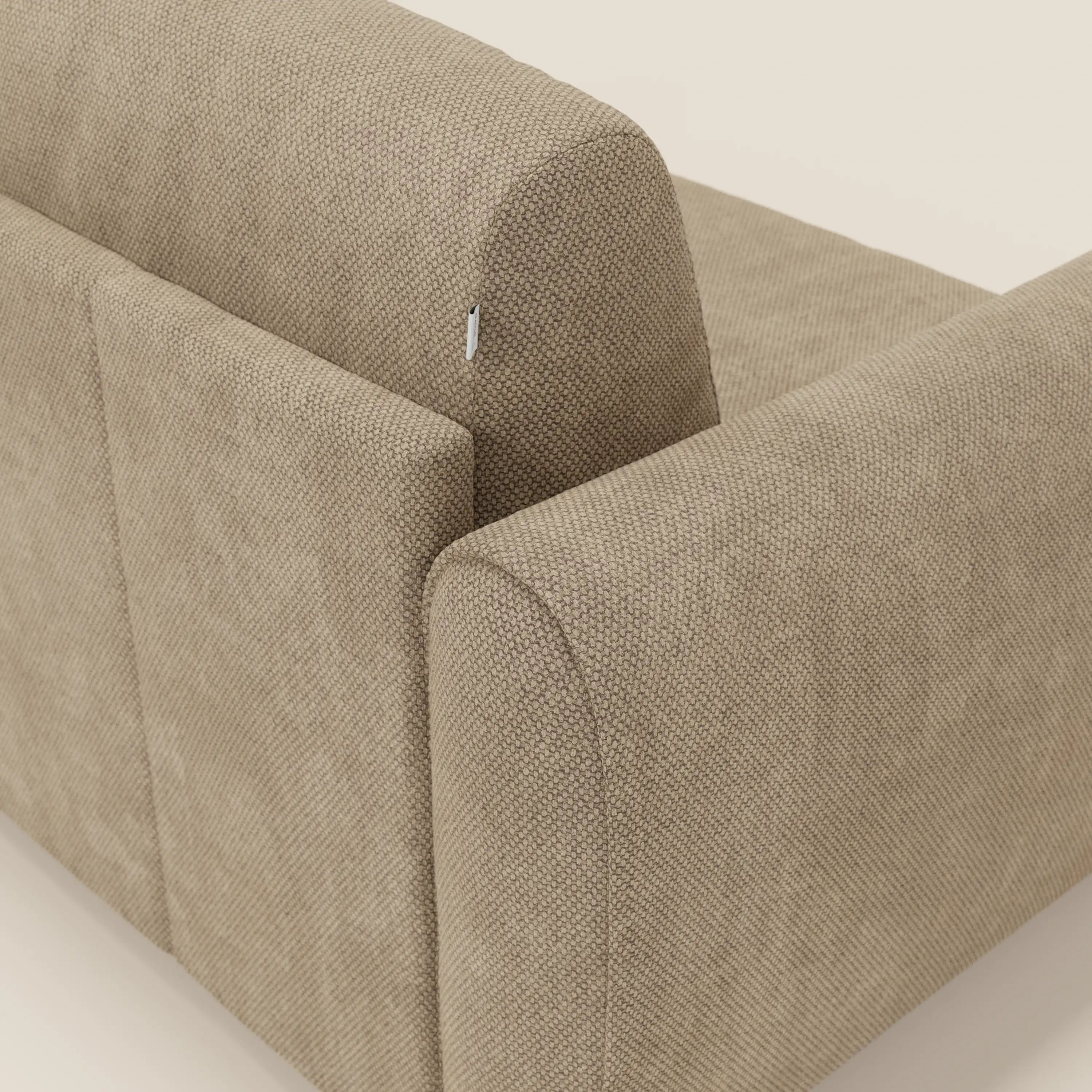 Un primo piano mostra lo schienale e il bracciolo di Kaleo Divano letto moderno di Divani.Store in tessuto impermeabile T26 beige con trama a rilievo e bordi arrotondati; è visibile una piccola targhetta bianca su uno sfondo chiaro.