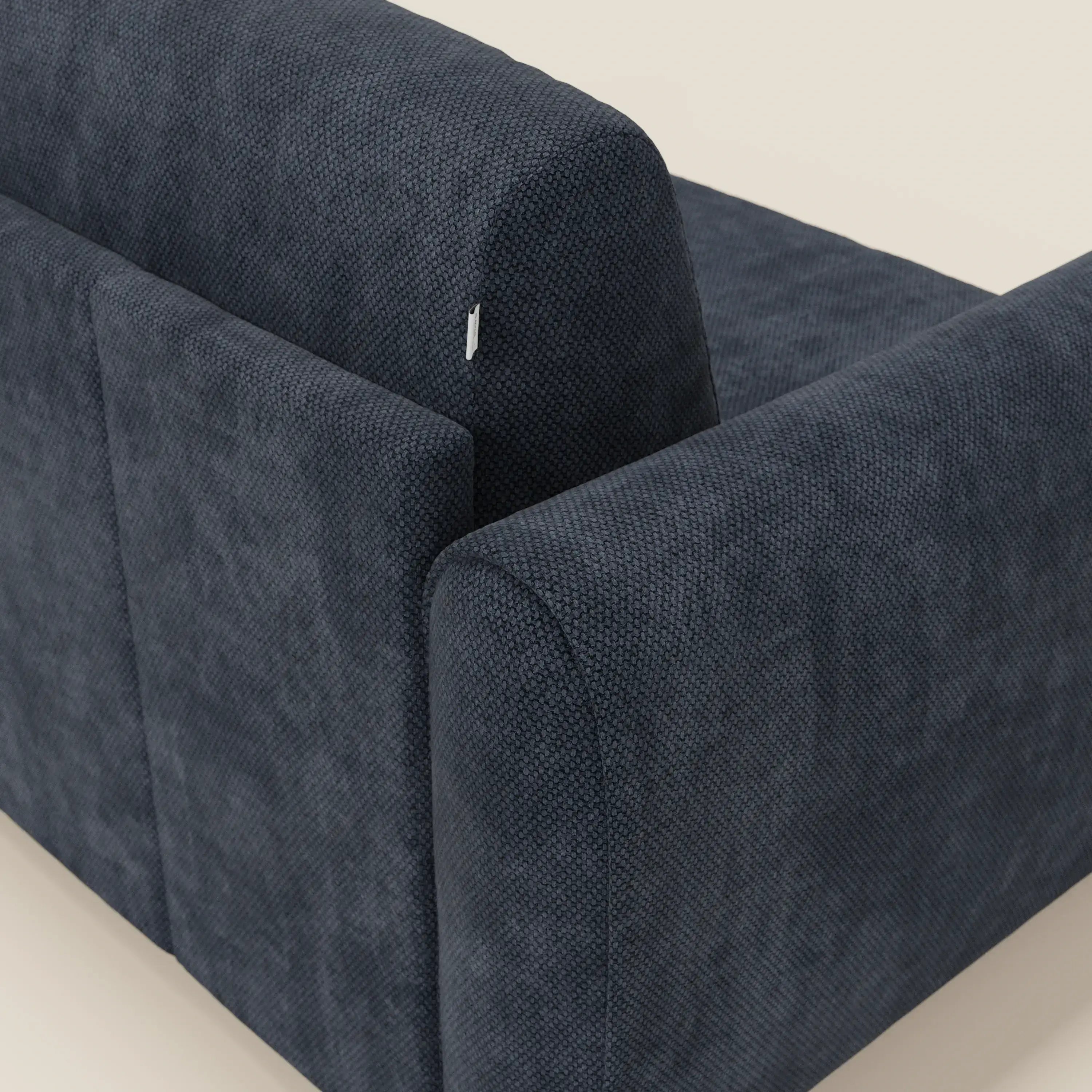 Un primo piano mostra lo schienale e il braccio di Kaleo Divano letto moderno di Divani.Store in tessuto impermeabile T26 blu navy, su uno sfondo beige chiaro. Una piccola etichetta in tessuto bianco vicino alla cucitura superiore ne accentua il design contemporaneo.