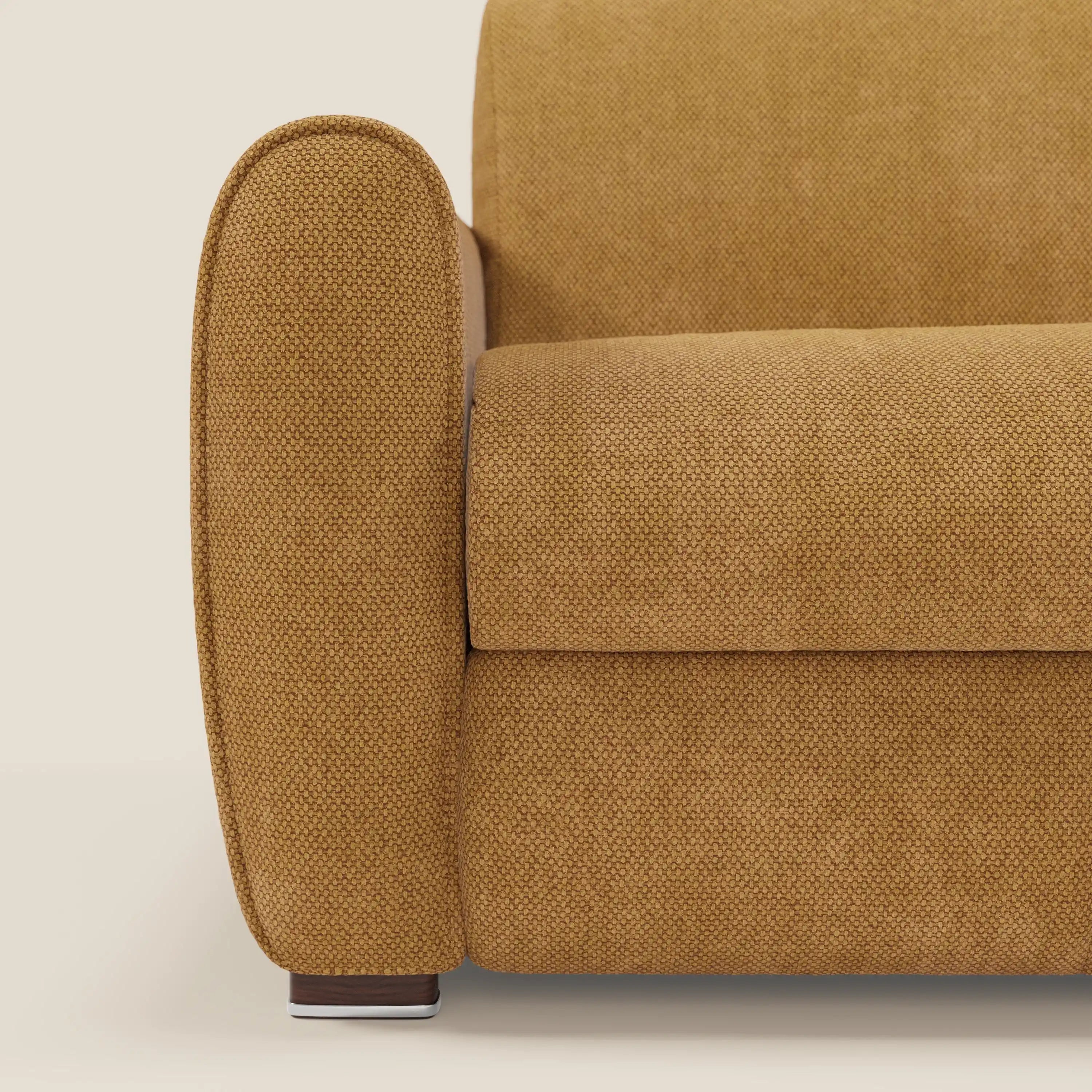 Primo piano di Kaleo Divano letto moderno di Divani.Store in tessuto impermeabile T26 giallo senape, che evidenzia il bracciolo sinistro arrotondato, il cuscino di seduta e la gamba corta in legno. Dispone di un materasso opzionale ad alta densità da 17 cm. Sfondo beige chiaro.