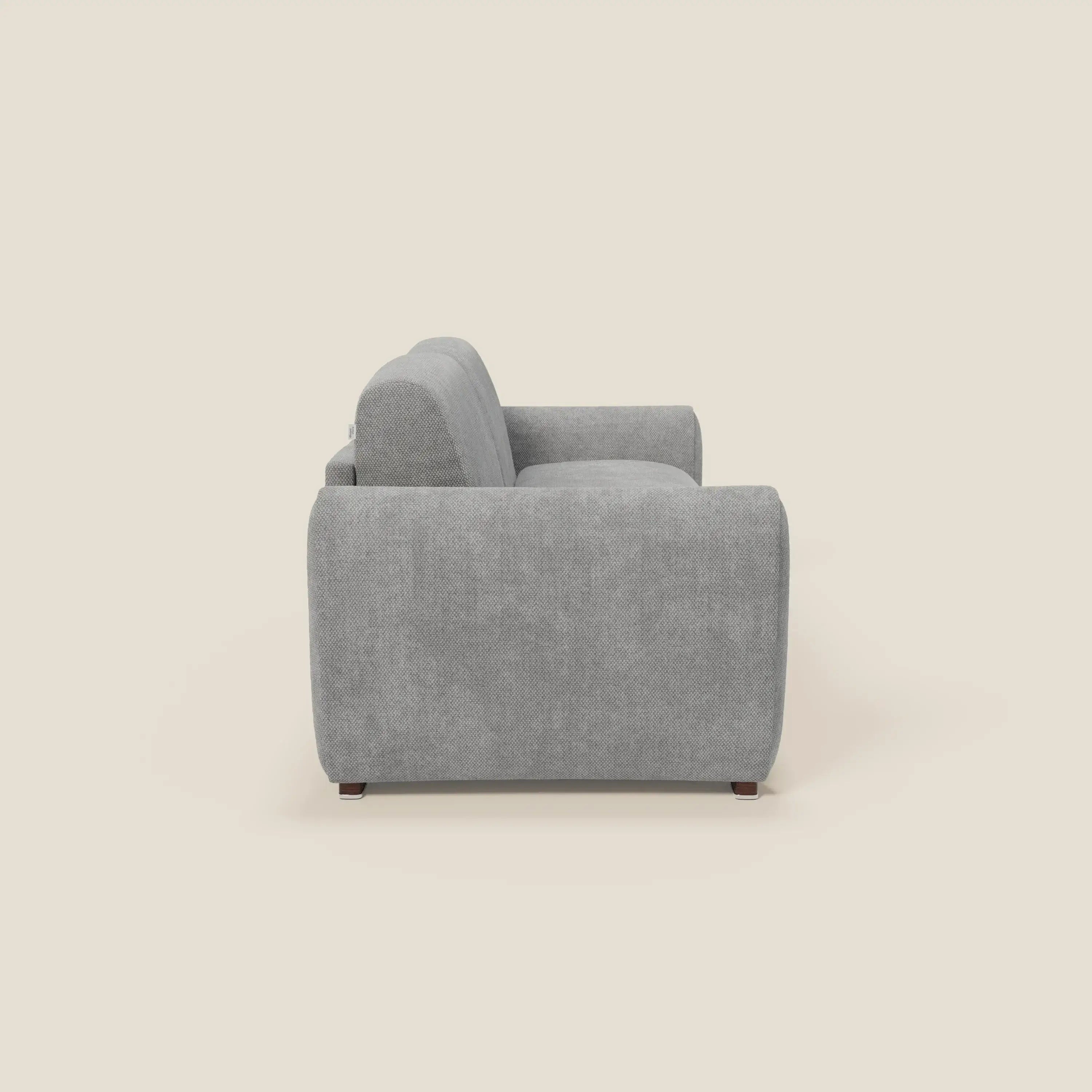 Il Kaleo Divano letto moderno di Divani.Store, in tessuto impermeabile grigio T26 con materasso ad alta densità da 17 cm, è presentato di lato su uno sfondo beige che mette in risalto lo spesso cuscino e le eleganti gambe in legno scuro.