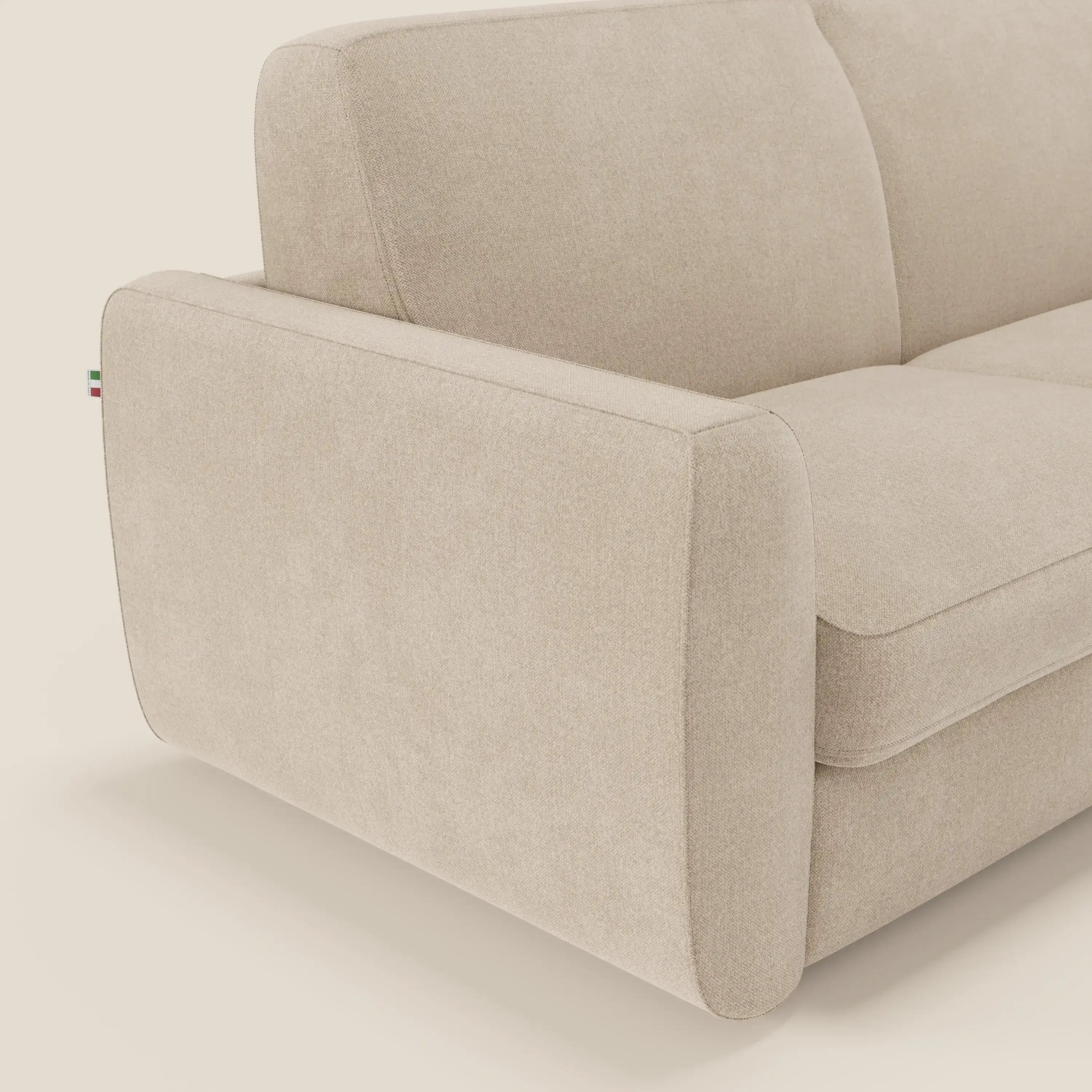Un primo piano del divano letto sfoderabile Leonida di Divani.Store in tessuto smacchiabile T23 beige mostra il bracciolo sinistro, la seduta, i cuscini dello schienale e una targhetta in tessuto verde, bianco e rosso nell'angolo posteriore.
