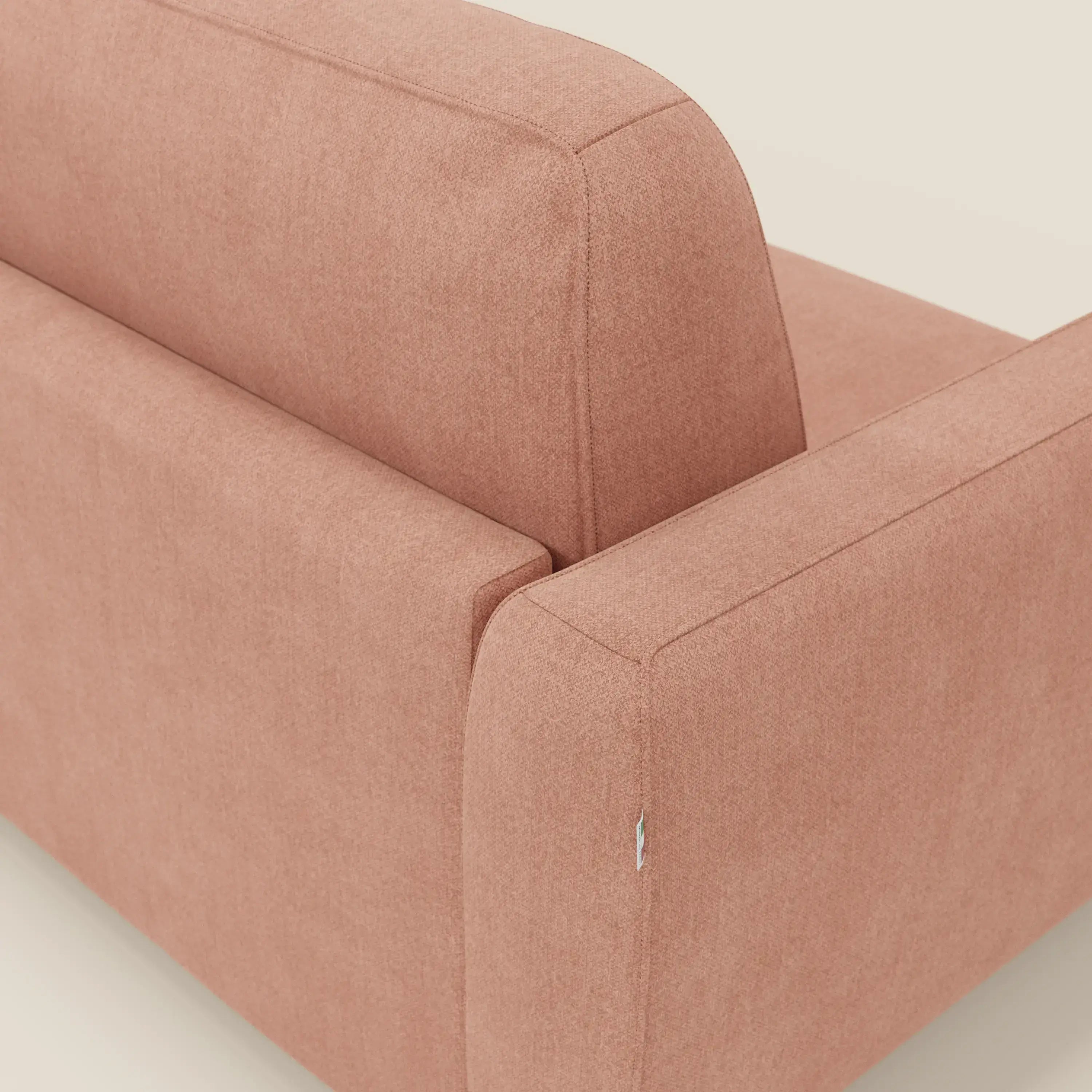 Un primo piano del divano letto sfoderabile Leonida di Divani.Store in tessuto smacchiabile T23 pesca mostra lo schienale, il bracciolo con cuciture a vista e la trama del tessuto, oltre a una piccola etichetta. Lo sfondo è chiaro e neutro.