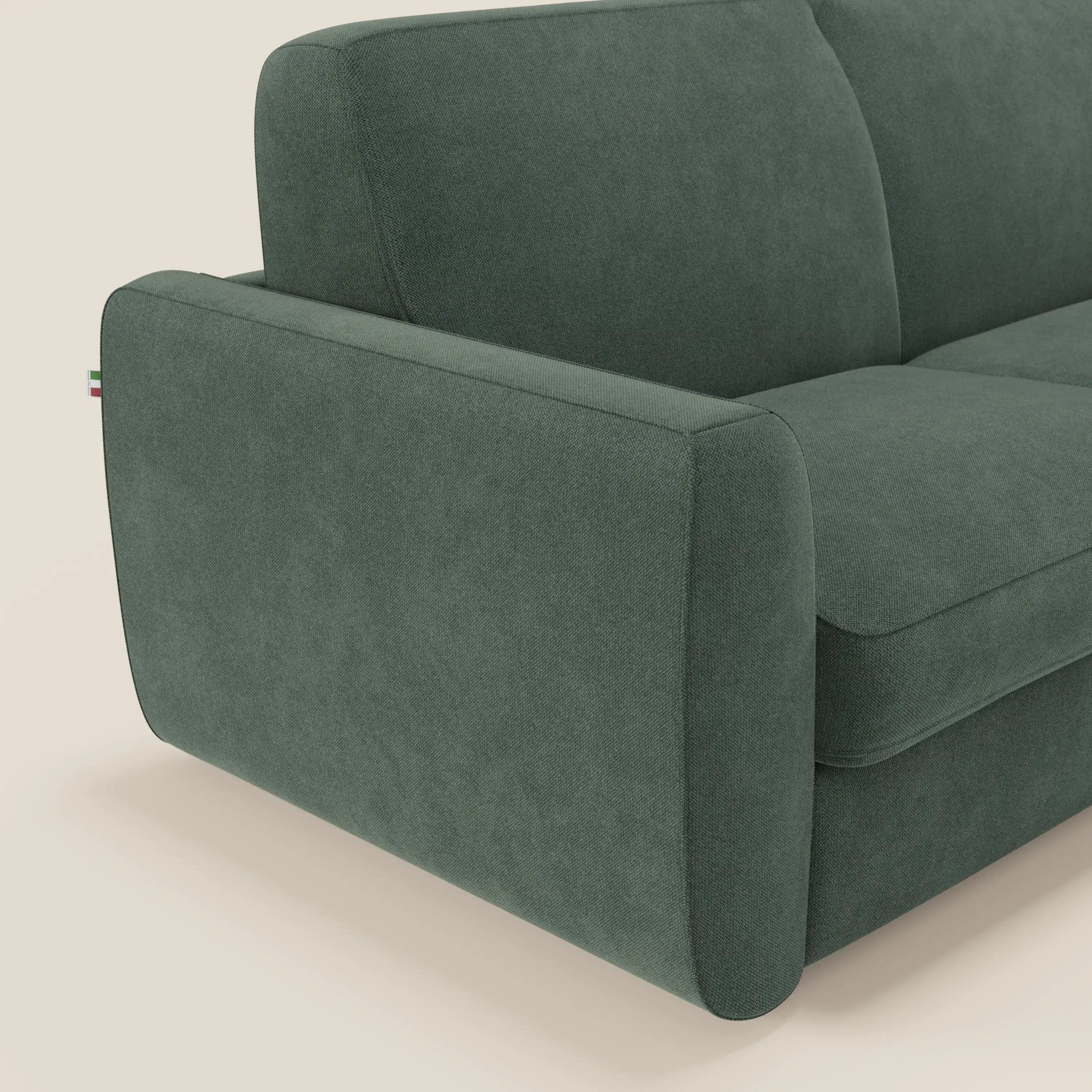 Un primo piano del divano letto sfoderabile Leonida di Divani.Store in smacchiabile T23 verde mostra il bracciolo sinistro con la targhetta della bandiera italiana e i morbidi cuscini scamosciati. Lo sfondo è bianco sporco. Dispone di materasso e cuscino H18 cm.