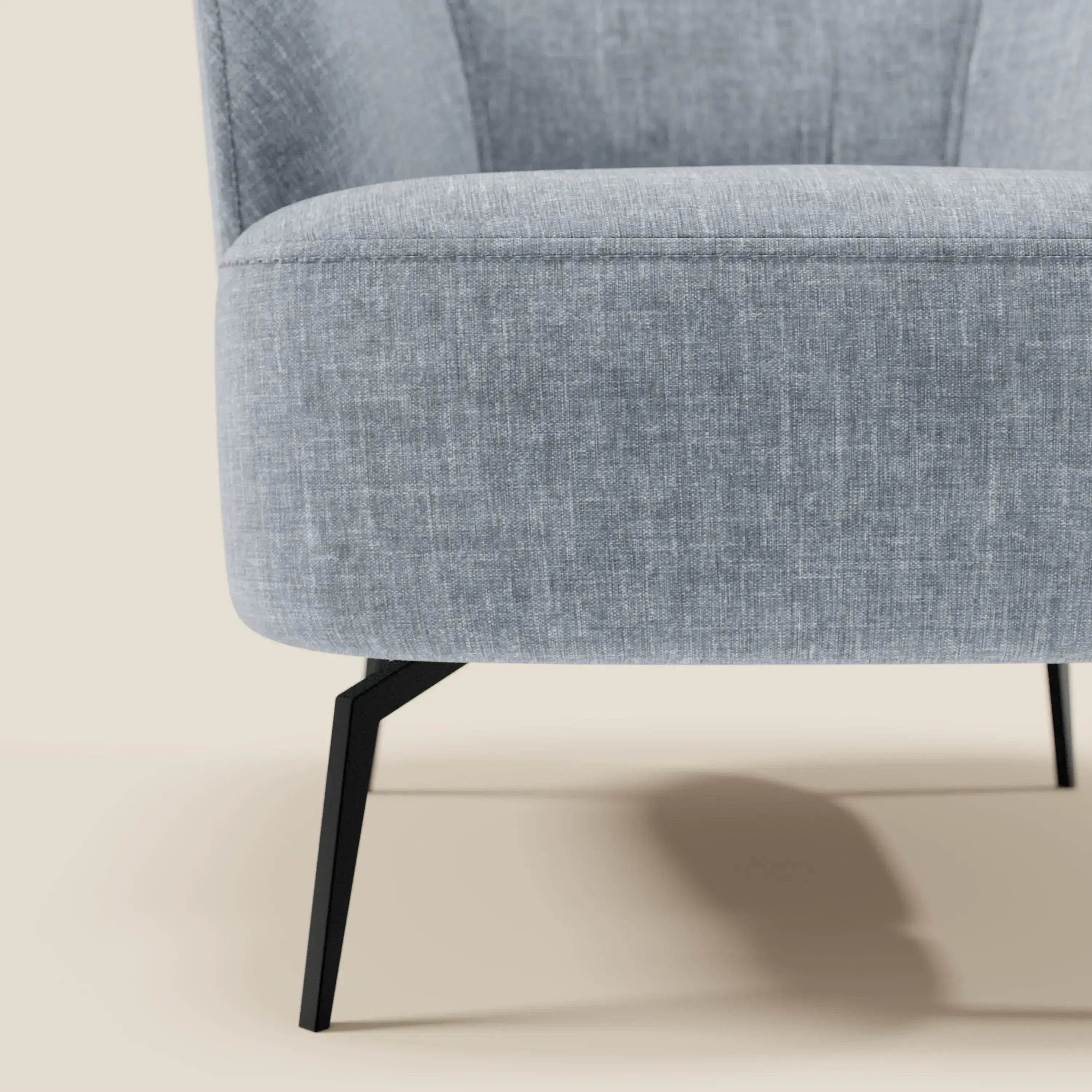 Primo piano della poltroncina Renoa di Divani.Store, caratterizzata da un elegante design moderno, dal comfort avvolgente e dal tessuto smacchiabile T24 carta da zucchero. Le linee pulite e le sottili gambe in metallo nero spiccano su uno sfondo beige.