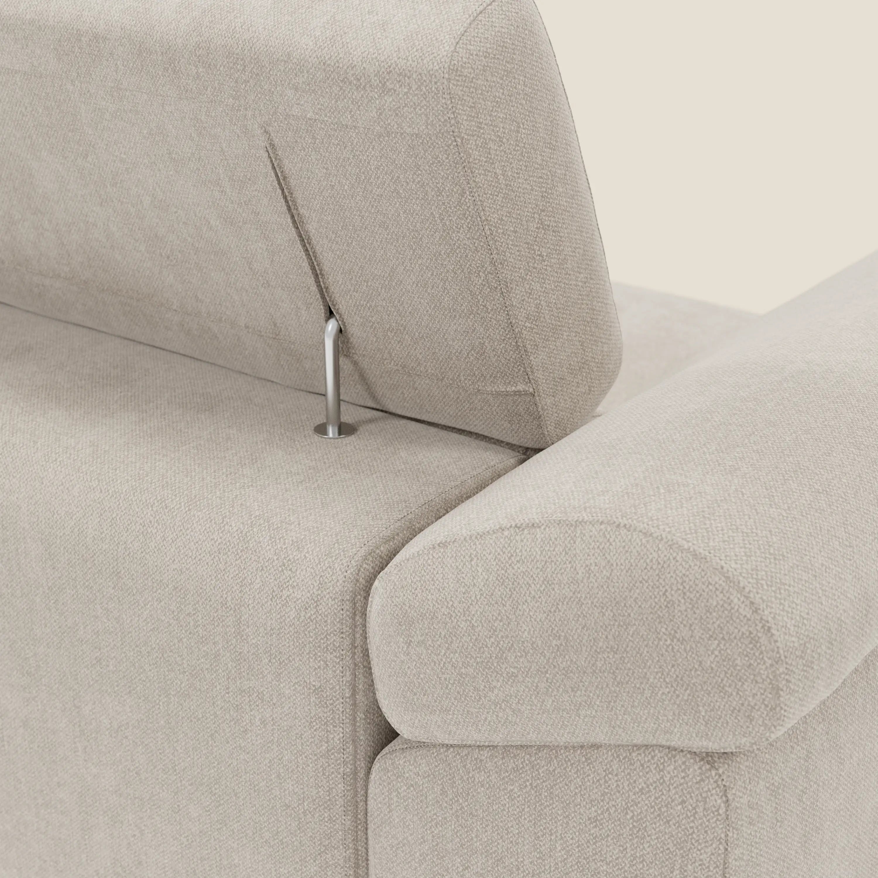 Primo piano del Divano angolare Donatello di Divani.Store in tessuto smacchiabile T23 panna, che mette in evidenza le sedute scorrevoli, i braccioli portaoggetti, il poggiatesta regolabile con asta metallica e il rivestimento in salvaspazio finemente intrecciato su uno sfondo bianco sporco.